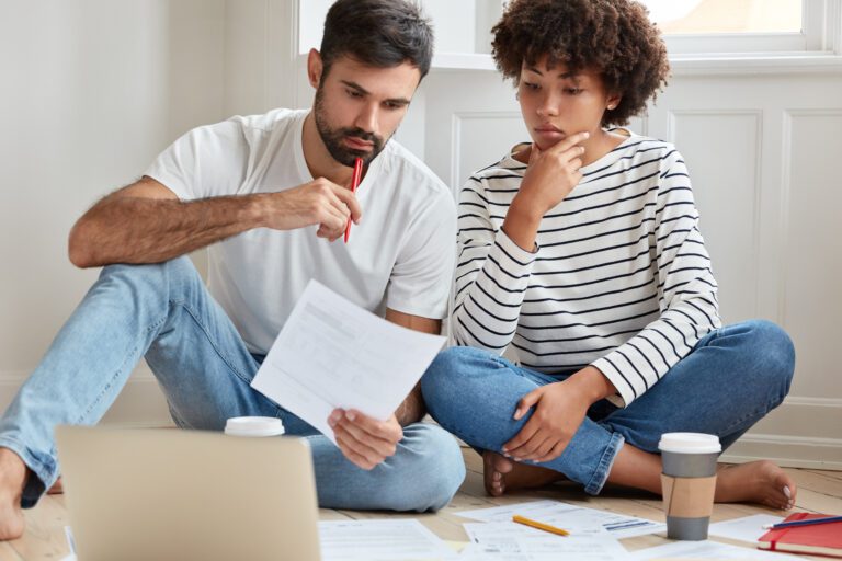 Indoor shot of serious mixed race woman and man study private arrangement, work from home, drink coffee, sit crossed legs on floor, read paper content attentively, try arrive at common conclusion